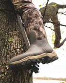 Person climbing a tree using a climbing harness and foot on a boot, with camouflage pants and a natural outdoor setting.