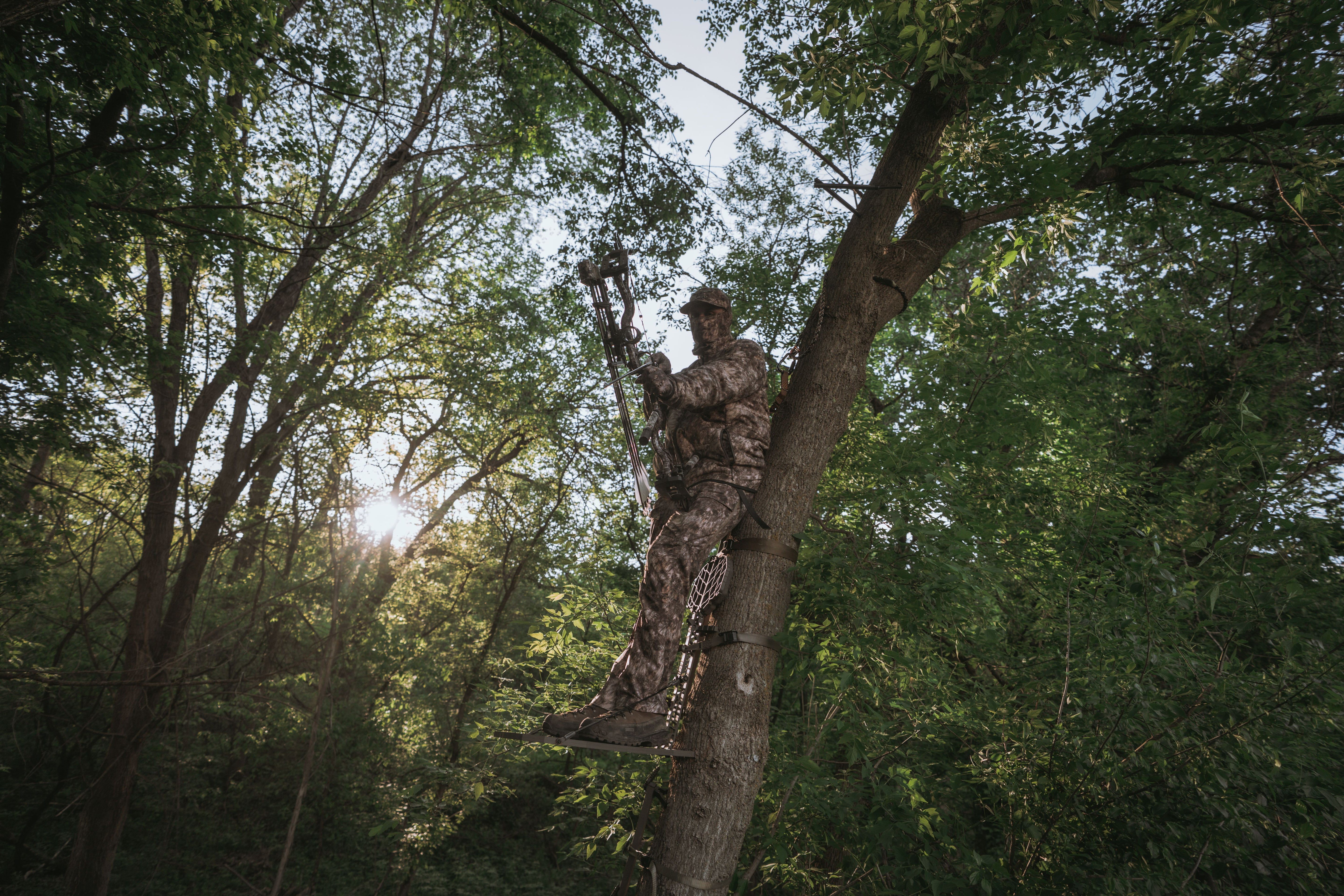 Bowhunter in RAPX camo gear standing in a treestand at sunrise, fully concealed in dense green timber—ideal for desktop header banner.