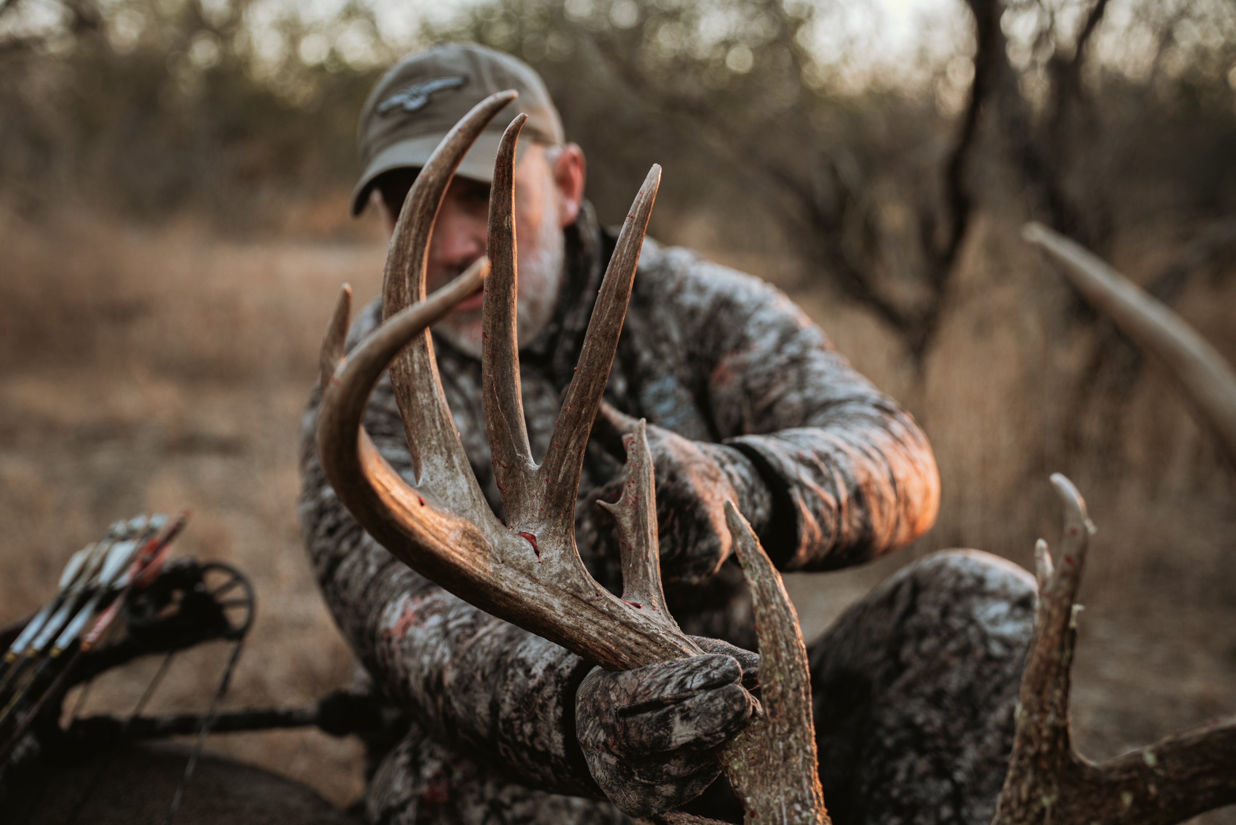 Bowhunter in ASIO RAPTOR camo grips mature whitetail antlers after a successful hunt, representing ASIO Gear’s mission to elevate the whitetail pursuit.
