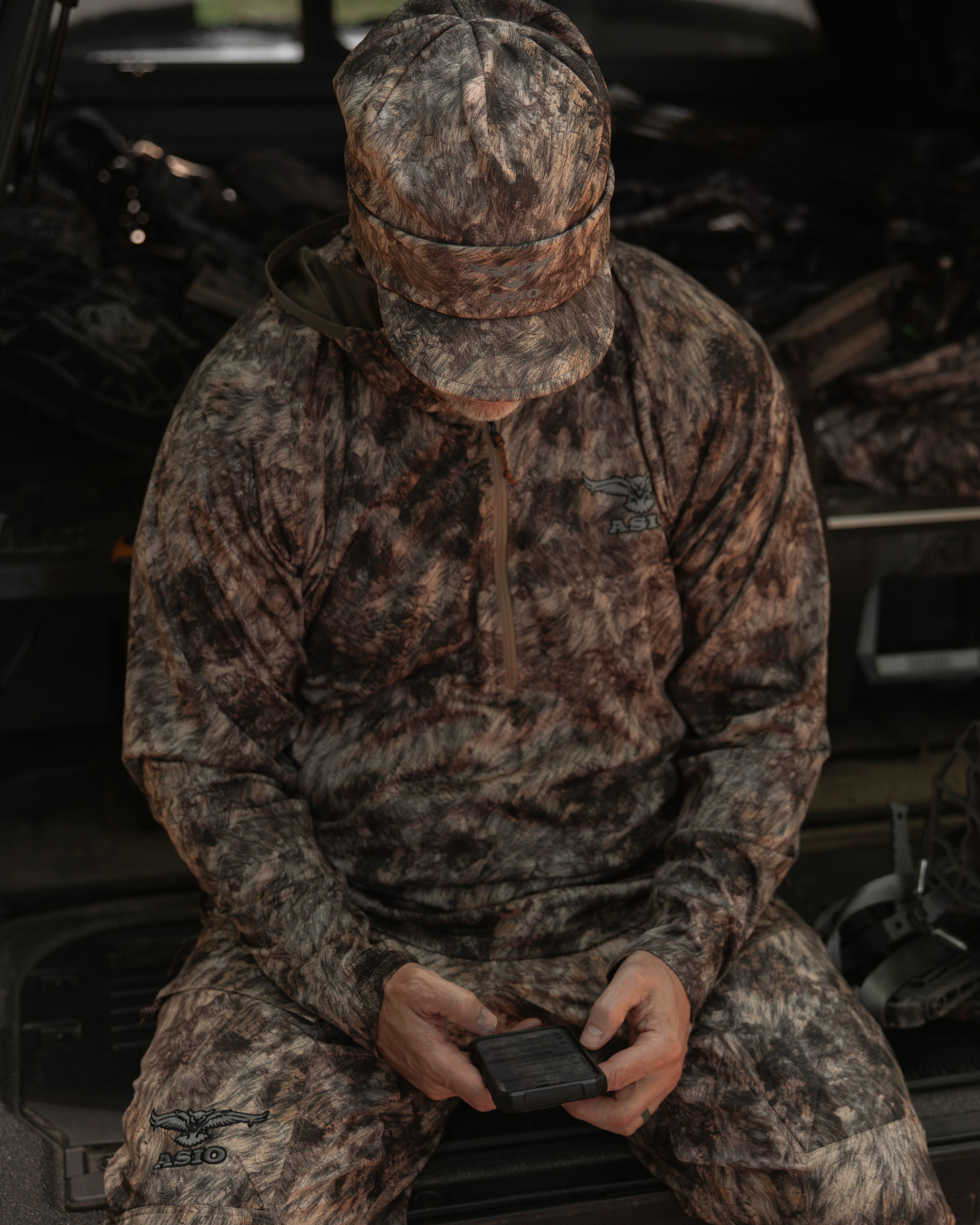 Hunter in RAPX camo fleece-lined billed beanie and full ASIO system sitting on tailgate with bow and gear in background